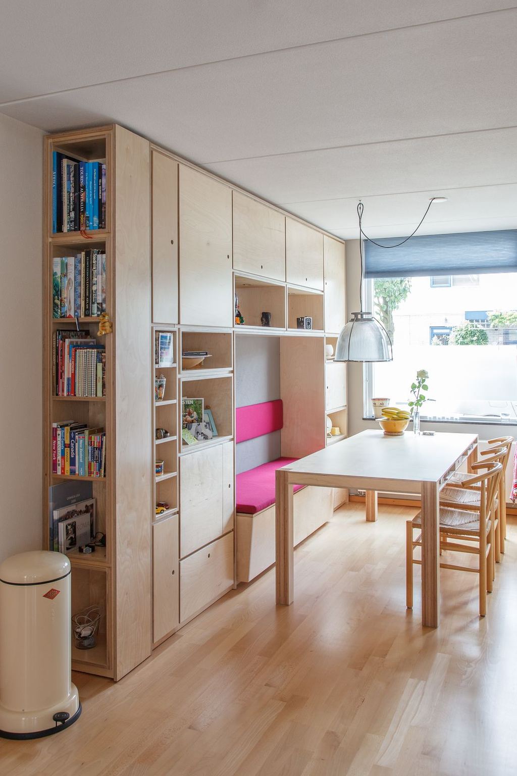 Wandvullend kastensysteem met een roze zitbank-nis, dat overgaat in een eettafel met vlechtstoelen.