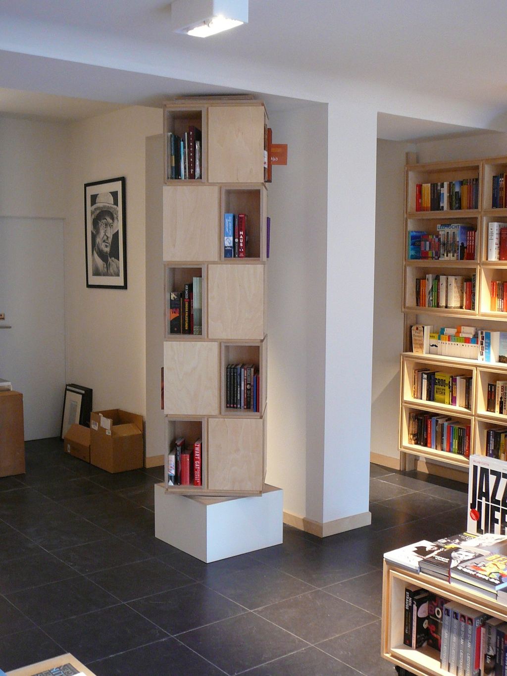 Smalle, torenvormige boekenkast van losse vakken, op een wit voetstuk.